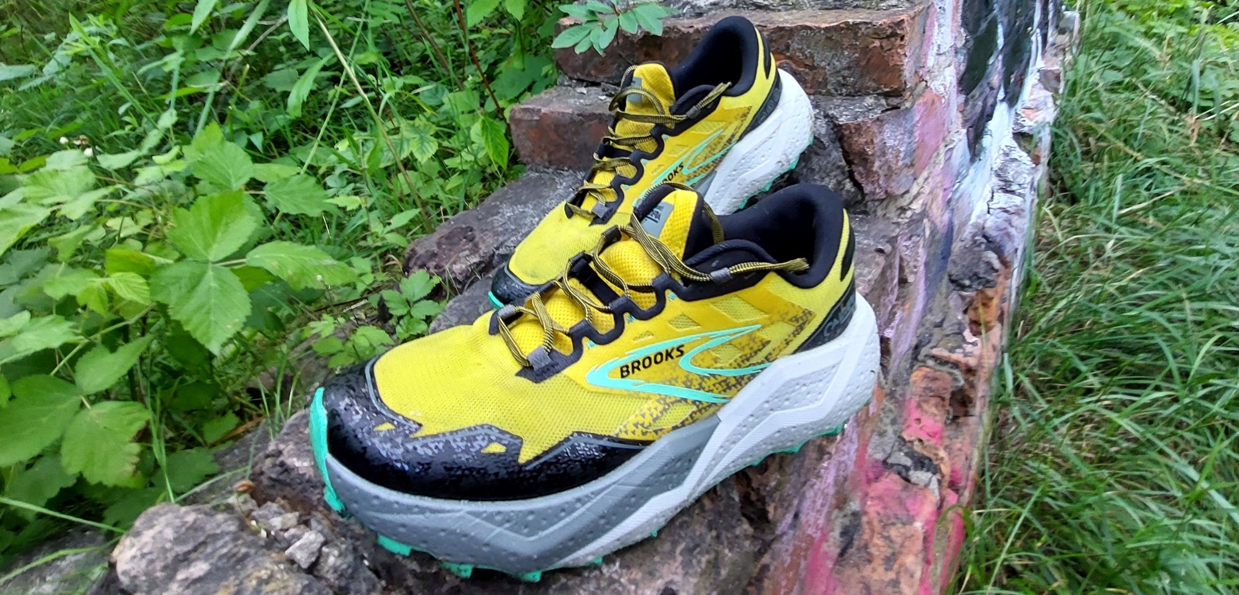 Żółte buty Brooks Caldera 7 leżace na fragmencie ukruszonego muru z czerwonej cegły. W tle zielona roślinność.